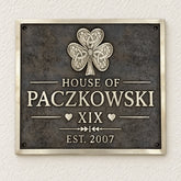 A rectangular metal plaque mounted on a textured off-white wall. The plaque has a dark textured background with a raised silver Celtic-style shamrock at the top and raised silver lettering reading 'HOUSE OF PACZKOWSKI', 'XIX', and 'EST.