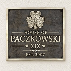 A rectangular metal plaque mounted on a textured off-white wall. The plaque has a dark textured background with a raised silver Celtic-style shamrock at the top and raised silver lettering reading 'HOUSE OF PACZKOWSKI', 'XIX', and 'EST.