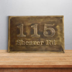 Brass house number sign with '115 Shearer Rd' on a wooden surface and light gray background