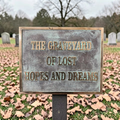 Bronze memorial sign with text 'The Graveyard of Lost Hopes and Dreams' in a cemetery.