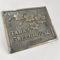 Bronze metal sign with floral design and text 'Tara's Greenhouse Est. 2025' on a white background