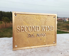 Gold plaque with 'SECOND WIND Est. 2020' text on a stone surface with a blurred natural background