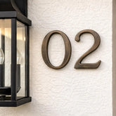 Bronze house numbers '02' on a textured white wall next to a black outdoor light fixture.