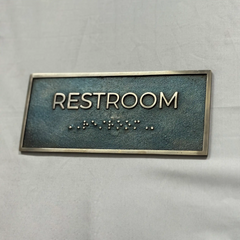 Rectangular sign with 'RESTROOM' text and braille on a gray background