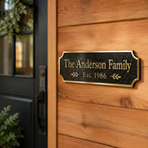 A personalized black metal front door sign with gold lettering that reads 'The Anderson Family Est. 1986', mounted on a warm wood-paneled wall next to a black door.