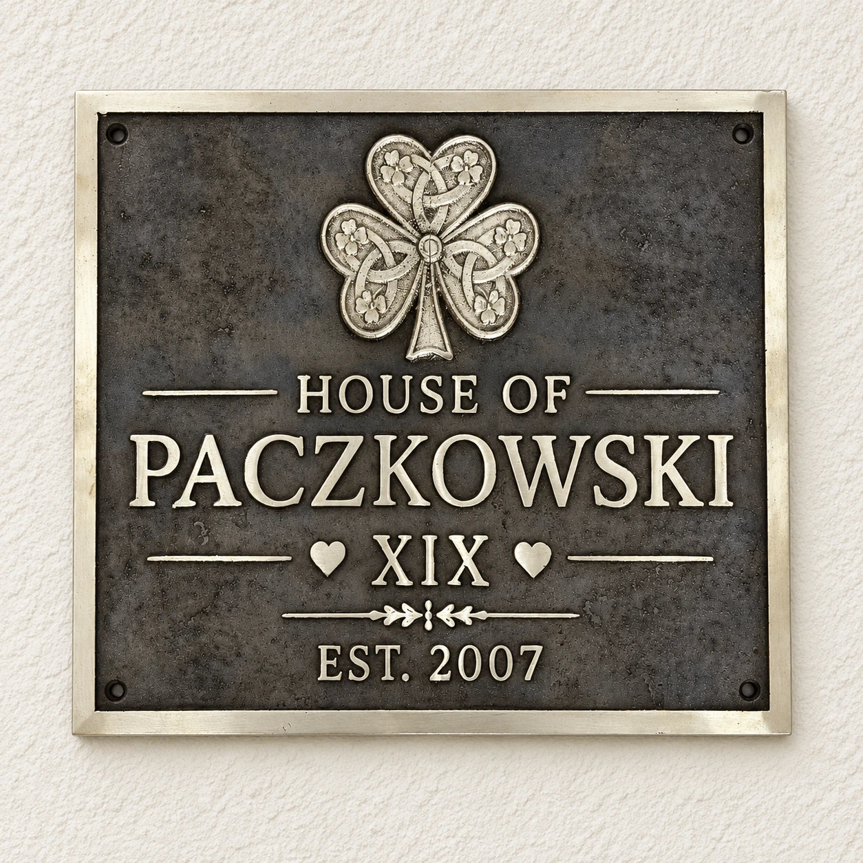 A rectangular metal plaque mounted on a textured off-white wall. The plaque has a dark textured background with a raised silver Celtic-style shamrock at the top and raised silver lettering reading 'HOUSE OF PACZKOWSKI', 'XIX', and 'EST.