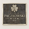 A rectangular metal plaque mounted on a textured off-white wall. The plaque has a dark textured background with a raised silver Celtic-style shamrock at the top and raised silver lettering reading 'HOUSE OF PACZKOWSKI', 'XIX', and 'EST.