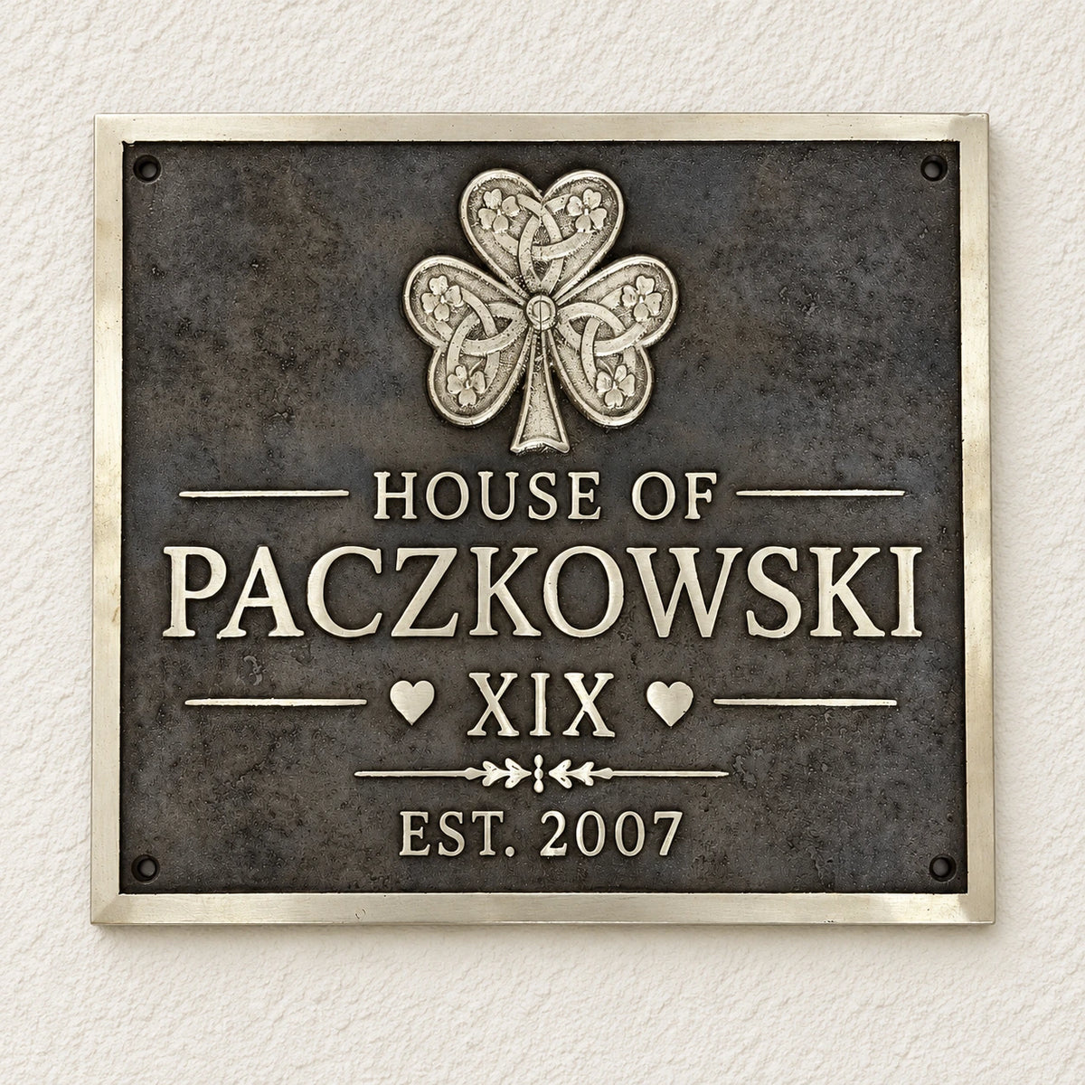 A rectangular metal plaque mounted on a textured off-white wall. The plaque has a dark textured background with a raised silver Celtic-style shamrock at the top and raised silver lettering reading 'HOUSE OF PACZKOWSKI', 'XIX', and 'EST.