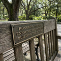Wooden bench with a bronze plaque reading 'Honoring the past, Enjoying the present, Building the future' in a park setting.