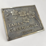 Bronze metal sign with floral design and text 'Tara's Greenhouse Est. 2025' on a white background