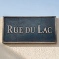 Bronze Street sign reading 'RUE DU LAC' on a textured wall with a clear blue sky.