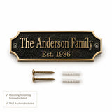 Custom family nameplate plaque with a black textured background and gold lettering, shown with two gold mounting screws and two white wall anchors on a white background. A small text box at the bottom left notes that matching mounting screws and wall anchors are included.