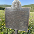 Bronze plaque with engraved text and emblem in a field of flowers and green hills.
