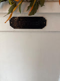 Decorative metal letter box sign on a white wall with green leaves above.