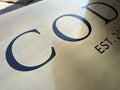 Brass sign with 'COD' and 'EST. 20' text on a reflective surface