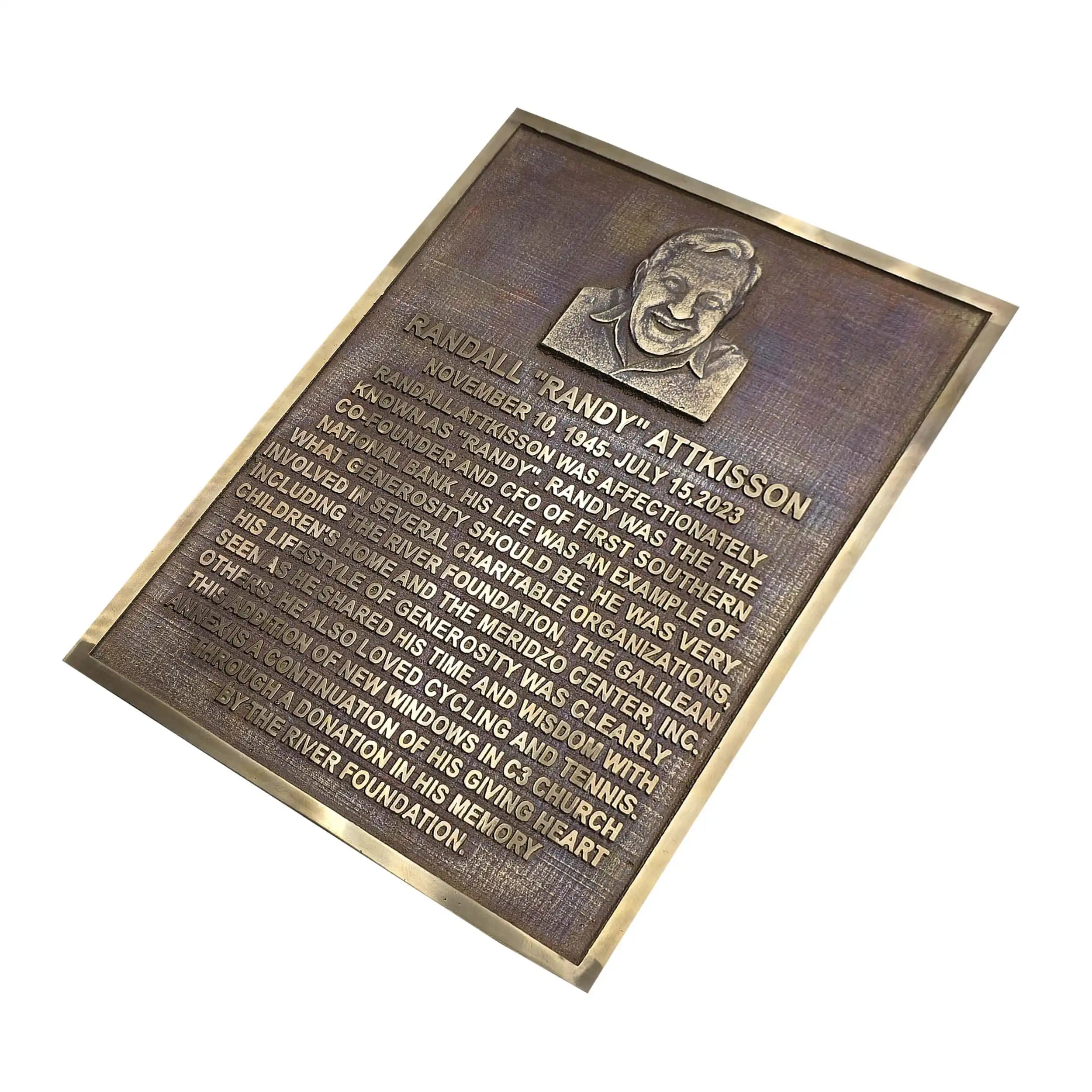 A rectangular bronze memorial plaque with a raised relief portrait at the top and engraved text below, shown at an angle on a plain white background.