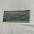 Rectangular sign with 'RESTROOM' text and braille on a gray background