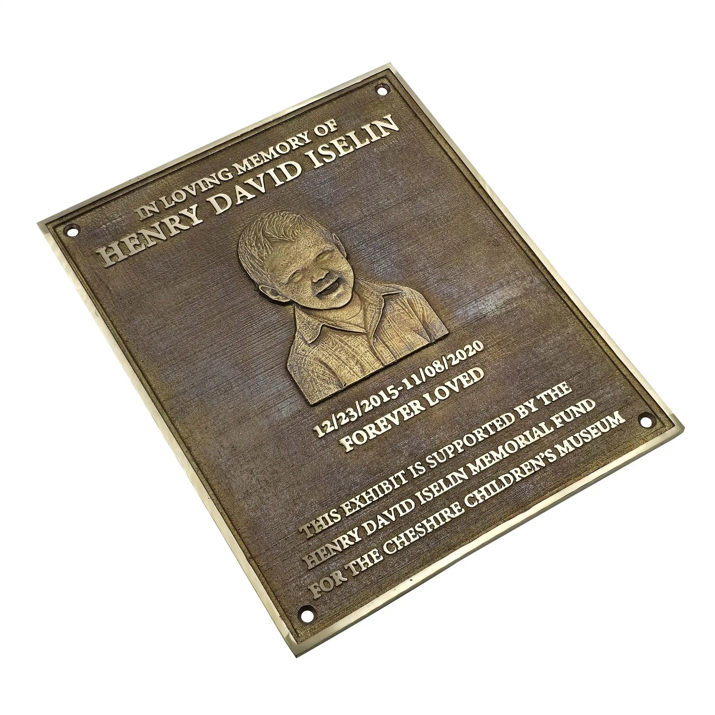 A rectangular bronze memorial plaque with rounded corners, featuring an engraved portrait of a young boy and text reading 'In Loving Memory of Henry David Iselin' and 'Forever Loved.' The plaque is photographed at an angle on a plain white background.
